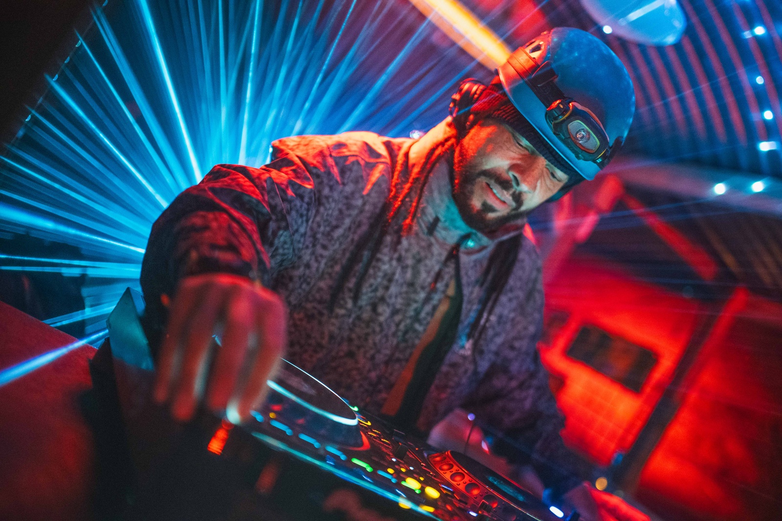 A DJ in a helmet and head torch at the decks, under blue lasers and red light.