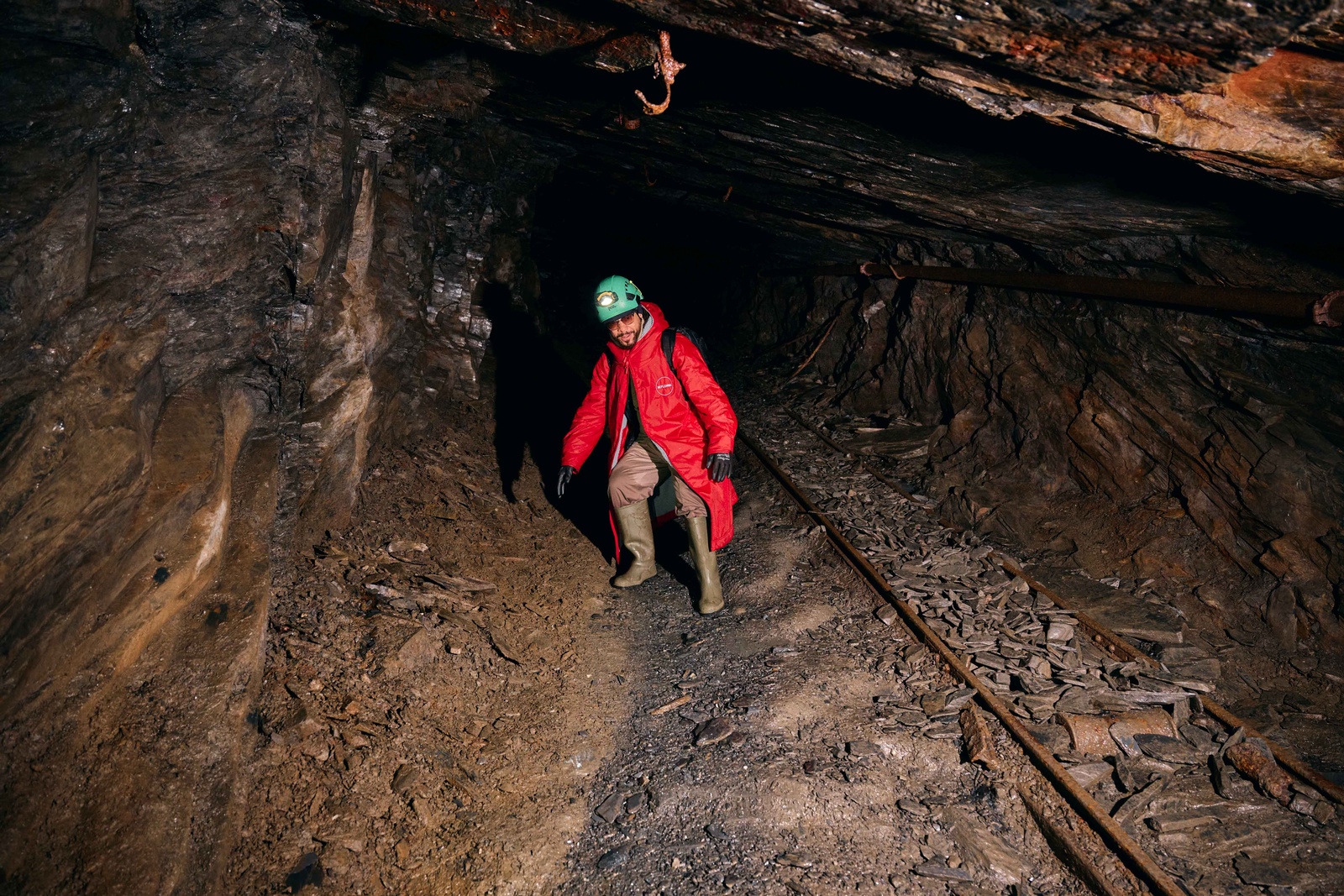 A person in a red coat, helmet and boots walking along old rails in a mine tunnel.